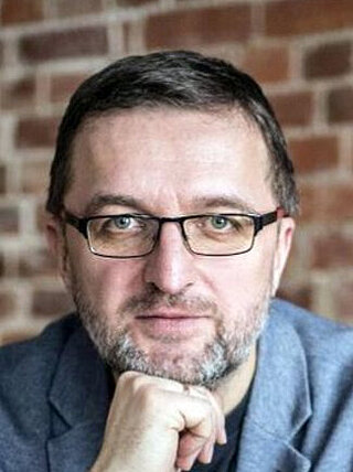 Uśmiechnięty mężczyzna w średnim wieku, o brązowych włosach, noszący okulary i błękitną marynarkę.