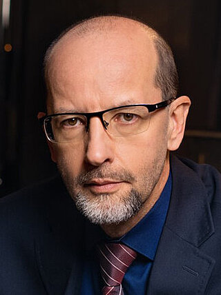 Poważny, starszy, łysiejący mężczyzna, noszący okulary z ciemnymi oprawkami. MA na sobie granatową marynarkę, niebieską koszulę i bordowy, prążkowany krawat.