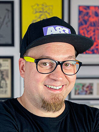 Szeroko uśmiechnięty młody mężczyzna noszący kolorowe plastikowe okulary, czapkę z daszkiem oraz kozią bródkę. 